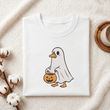 Folded white cotton t-shirt mockup featuring a cute duck in a ghost costume holding a trick-or-treat bucket machine embroidery design, styled with cotton stems and a wooden cup