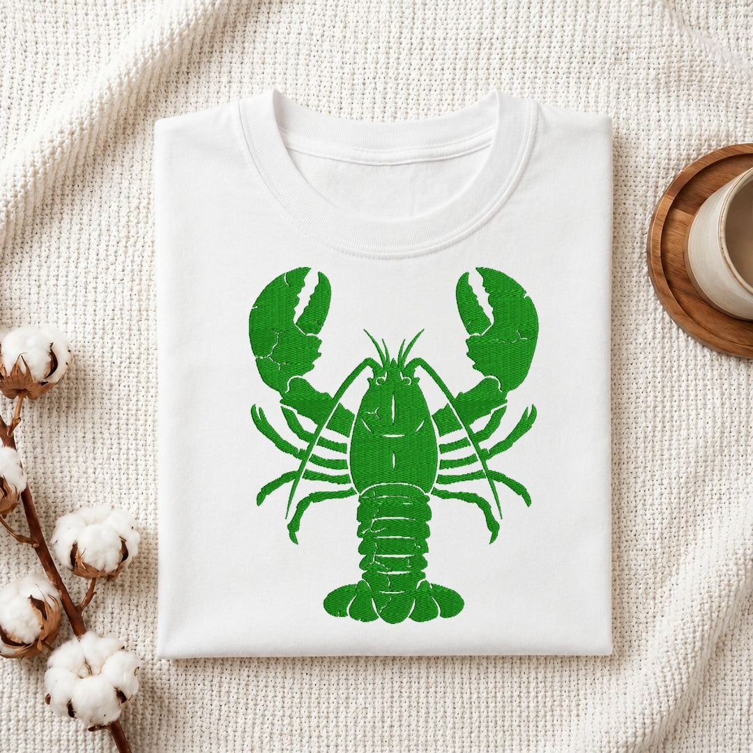 A folded white crewneck t-shirt featuring a distressed green lobster graphic on the front, styled on a textured cream fabric background with cotton bolls and a wooden cup.