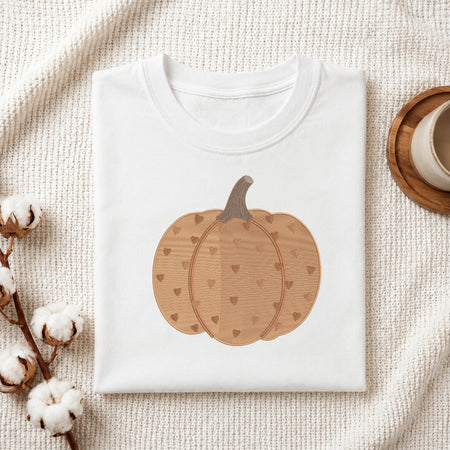 A folded white crewneck t-shirt featuring a graphic of a light brown pumpkin with a heart pattern and a gray stem, styled on a textured cream fabric with cotton stems and a wooden cup.