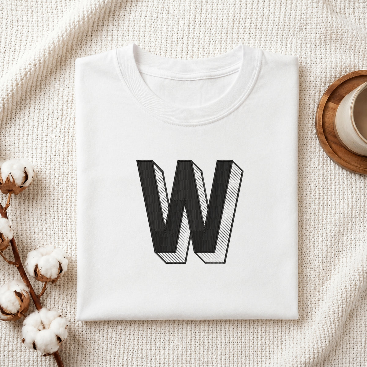 A folded white crewneck t-shirt with a large black block letter 'W' printed on the front, styled on a textured cream knit blanket with cotton bolls and a cup of coffee in the background.