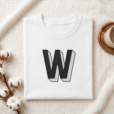 A folded white crewneck t-shirt with a large black block letter 'W' printed on the front, styled on a textured cream knit blanket with cotton bolls and a cup of coffee in the background.