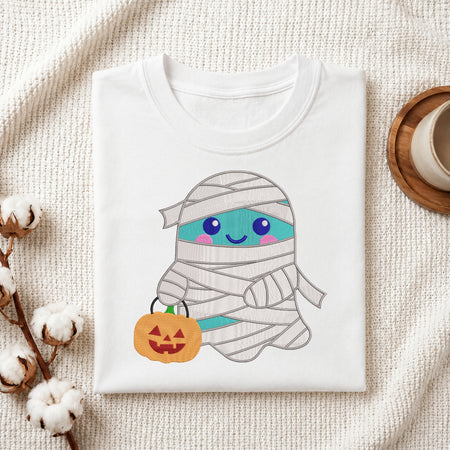 A folded white t-shirt with a cute cartoon mummy graphic, styled in a flat-lay on a textured cream fabric with cotton bolls and a cup on a wooden tray.