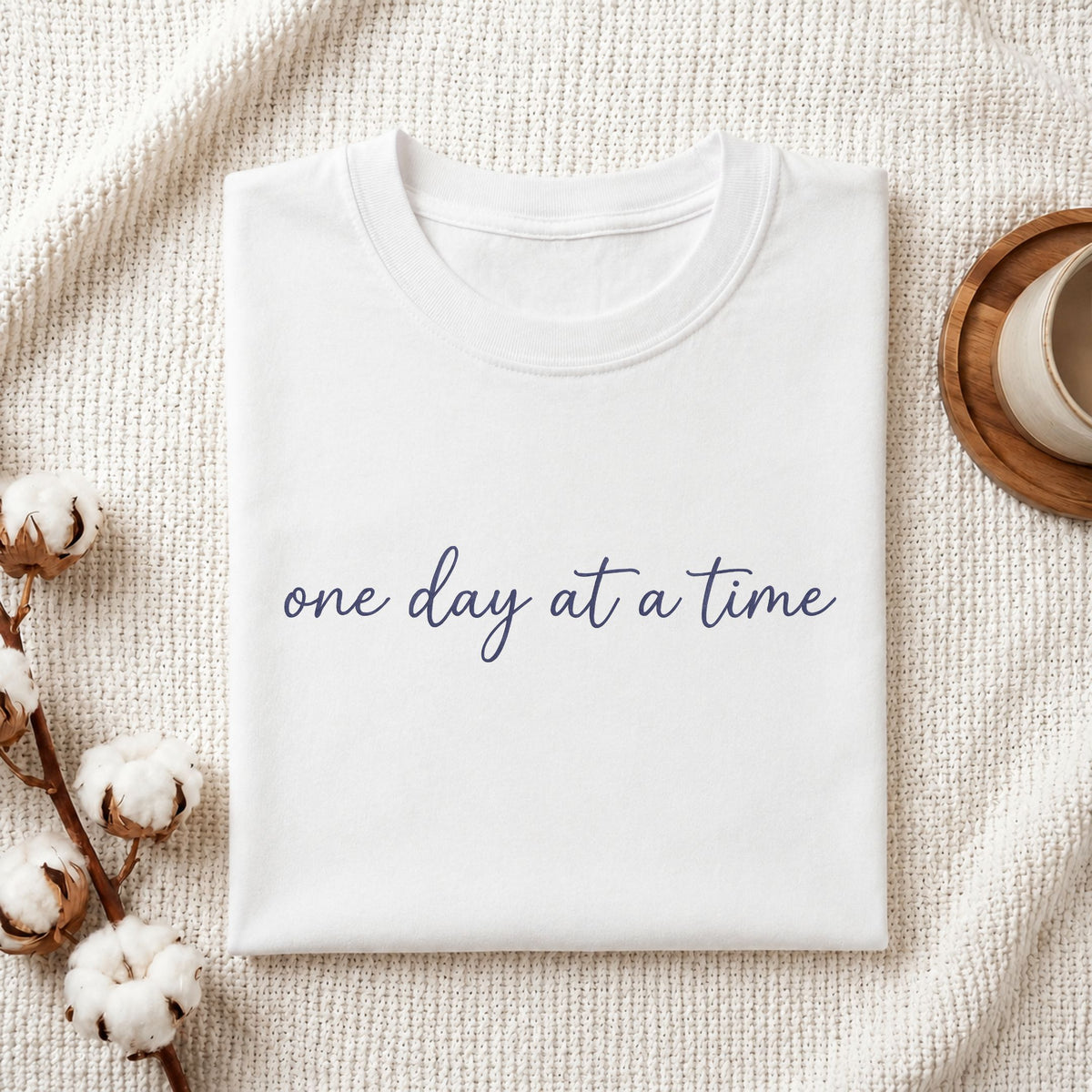 A folded white crewneck t-shirt with the phrase 'one day at a time' printed in navy blue script across the chest, styled on a textured cream knit blanket with cotton bolls and a cup in the background.