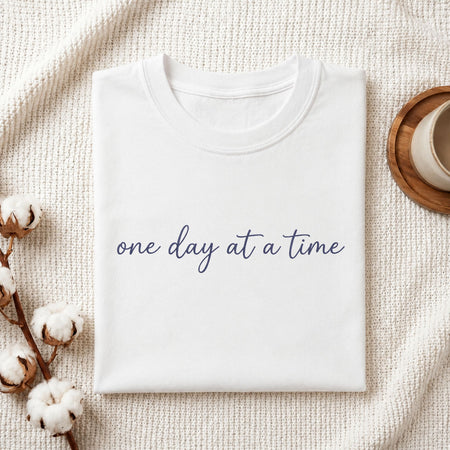 A folded white crewneck t-shirt with the phrase 'one day at a time' printed in navy blue script across the chest, styled on a textured cream knit blanket with cotton bolls and a cup in the background.