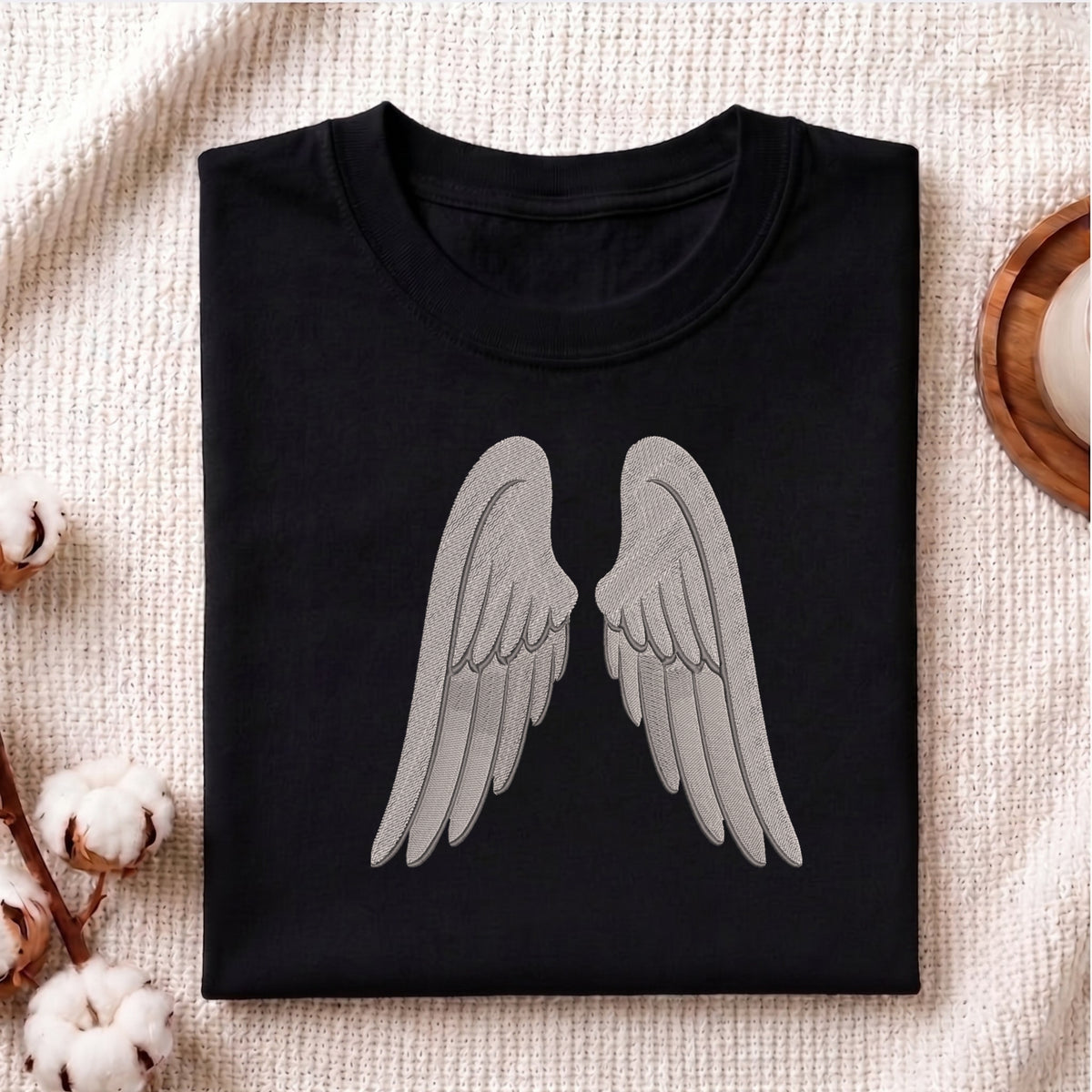 A folded black crewneck t-shirt with a large gray angel wings graphic on the front, displayed on a textured beige fabric background with cotton bolls and a wooden tray.