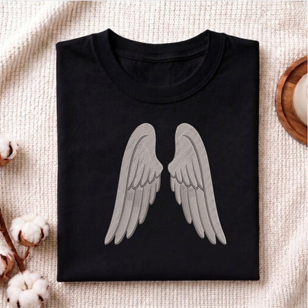 A folded black crewneck t-shirt with a large gray angel wings graphic on the front, displayed on a textured beige fabric background with cotton bolls and a wooden tray.