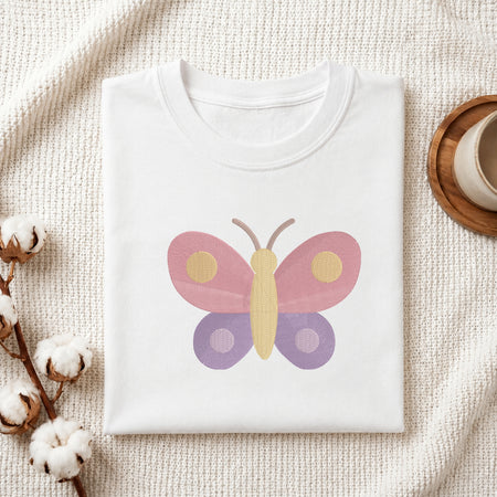 A folded white crewneck t-shirt with a colorful butterfly graphic on the front, styled on a textured cream knit blanket with cotton bolls and a wooden cup in the background.