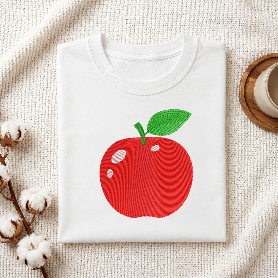A folded white crewneck t-shirt featuring a graphic of a red apple with a green leaf, styled on a textured cream fabric with cotton bolls and a wooden cup in the background.