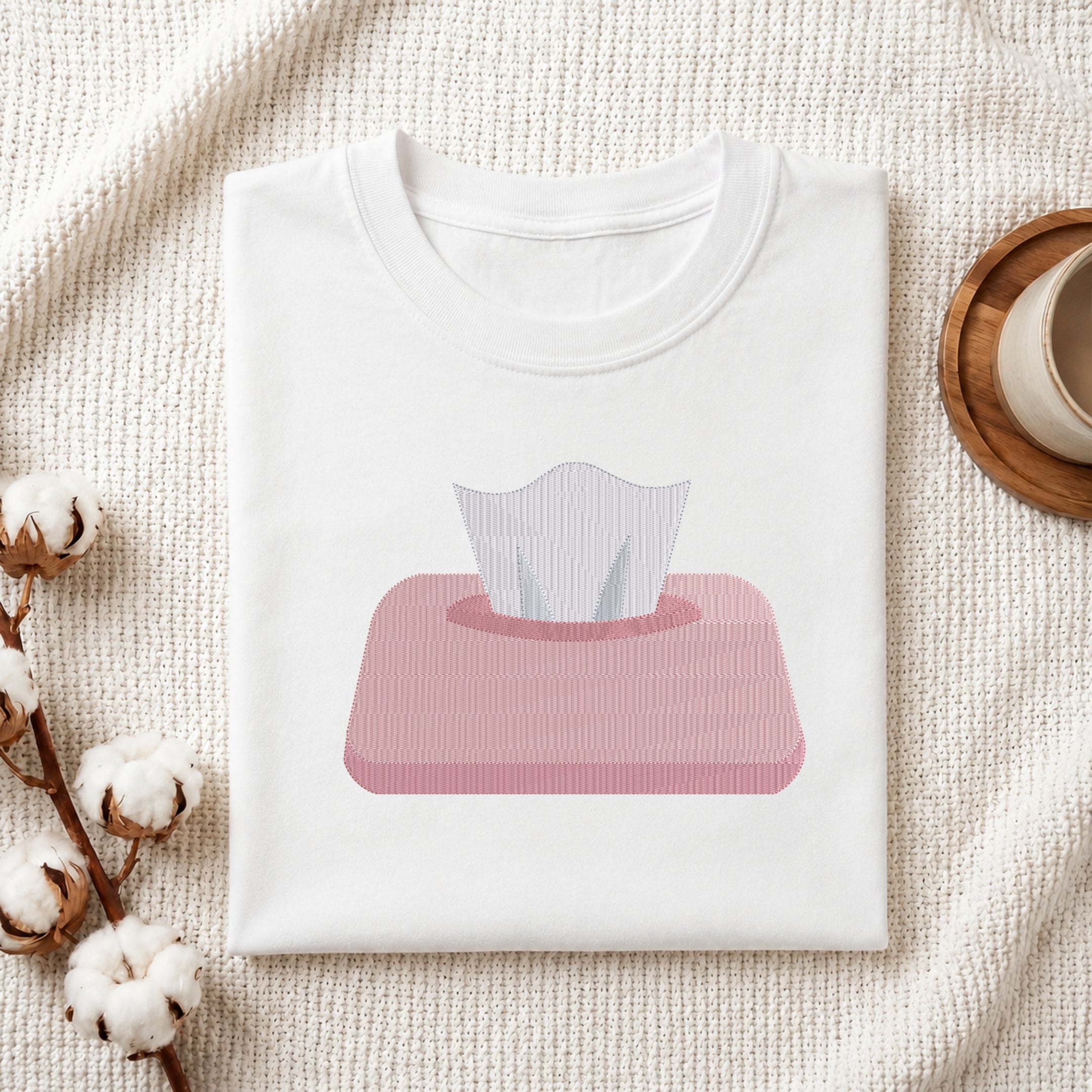 A folded white crewneck t-shirt with a pink tissue box graphic on the front, styled on a textured cream knit blanket with cotton bolls and a wooden cup in the background.