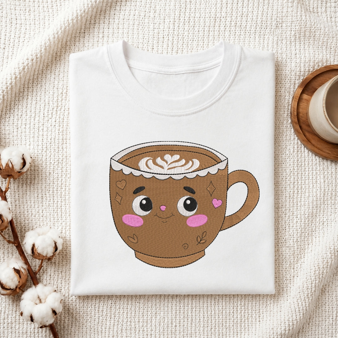 A folded white crewneck t-shirt featuring a cute cartoon coffee cup graphic, styled on a textured cream fabric with cotton stems and a wooden coaster.