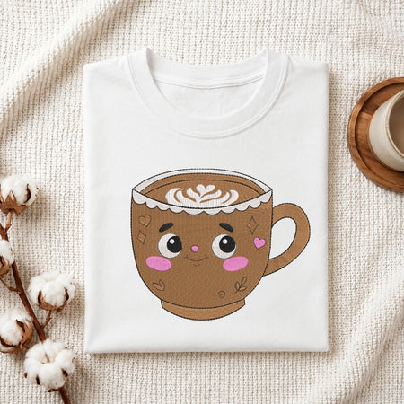 A folded white crewneck t-shirt featuring a cute cartoon coffee cup graphic, styled on a textured cream fabric with cotton stems and a wooden coaster.