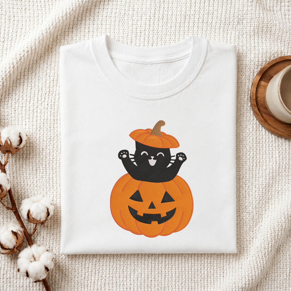Folded white cotton t-shirt mockup featuring a cute black cat wearing a pumpkin hat sitting inside a jack-o-lantern machine embroidery design, styled with cotton stems and a wooden cup