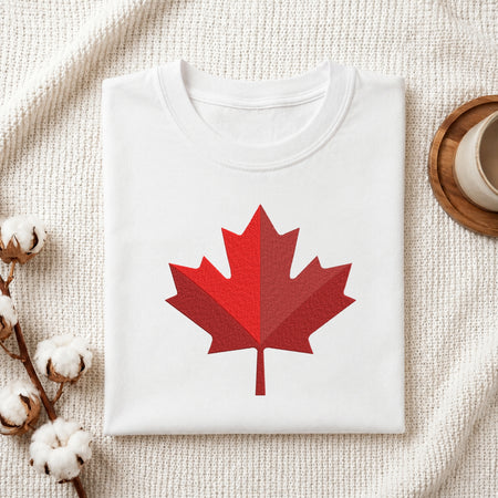 A folded white crewneck t-shirt with a red geometric maple leaf graphic on the front, styled on a textured cream knit blanket with cotton bolls and a wooden cup in the background.