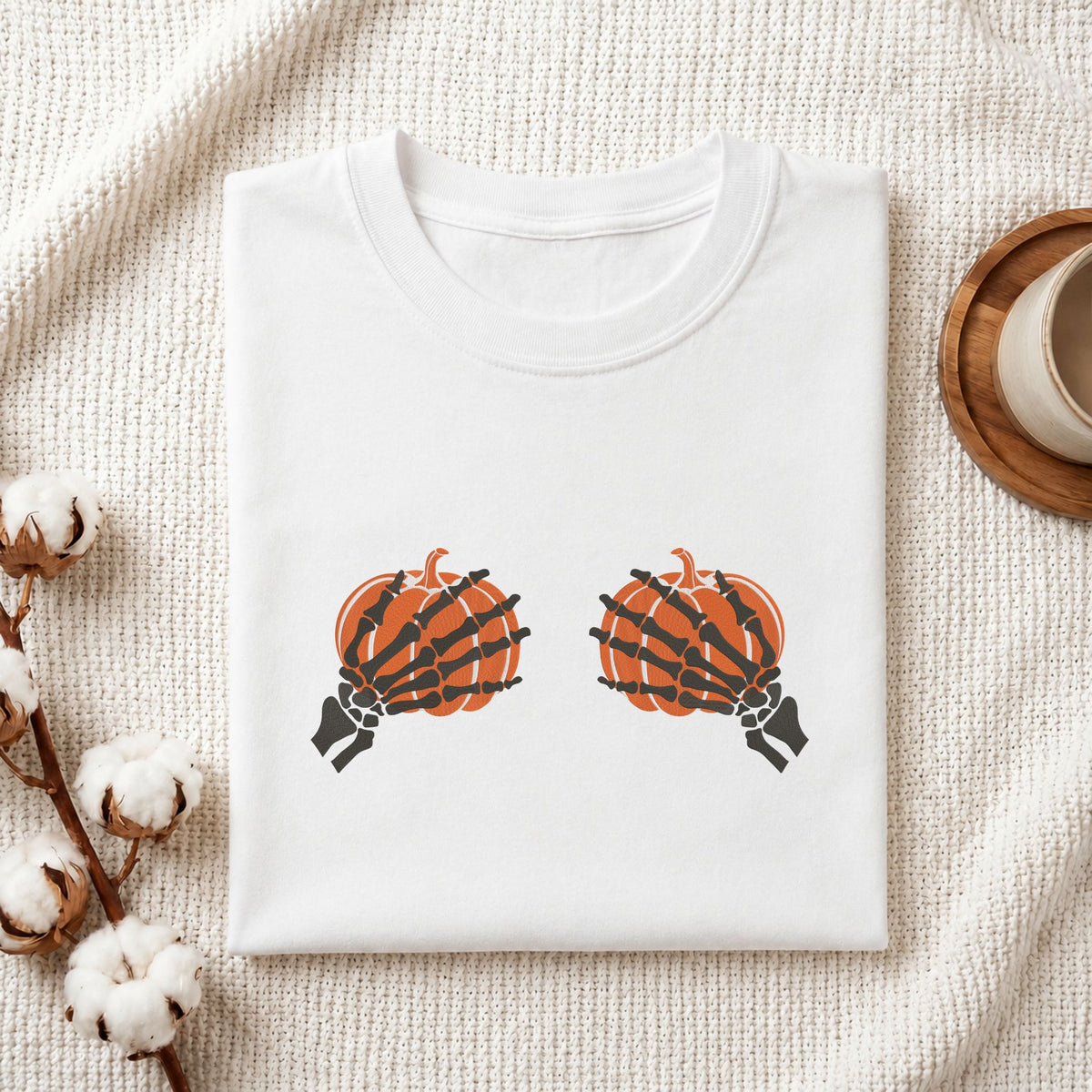  Folded white cotton t-shirt mockup featuring a skeleton hands holding pumpkins machine embroidery design, styled with cotton stems and a wooden cup   