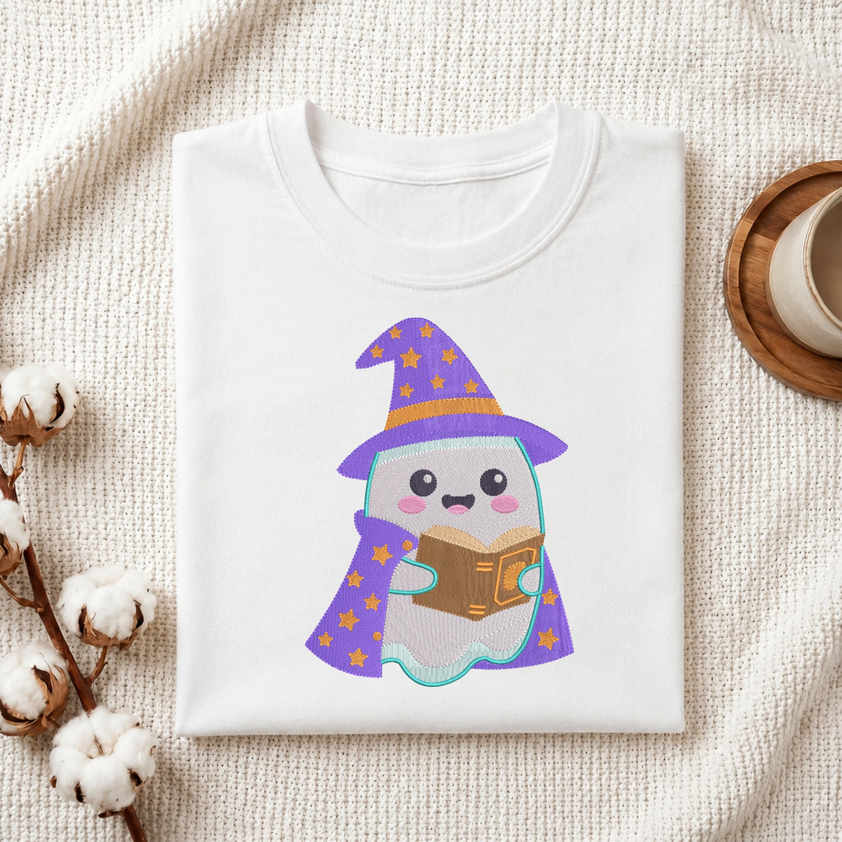 A folded white t-shirt with a cute cartoon ghost wearing a purple wizard hat and cape, reading a brown book. The t-shirt is styled in a flat-lay on a textured cream fabric with cotton stems and a wooden cup on a coaster.