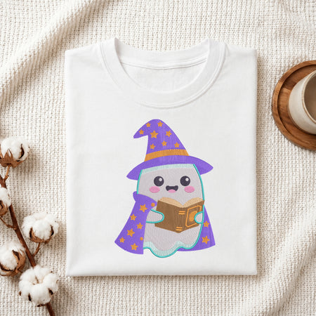 A folded white t-shirt with a cute cartoon ghost wearing a purple wizard hat and cape, reading a brown book. The t-shirt is styled in a flat-lay on a textured cream fabric with cotton stems and a wooden cup on a coaster.