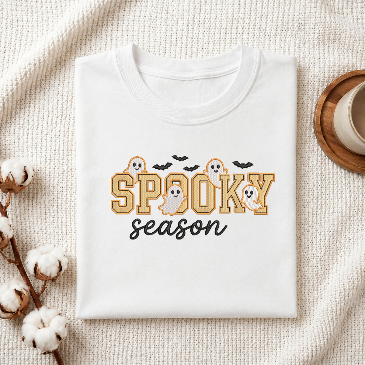A folded white t-shirt with a 'Spooky Season' graphic print, styled in a flat-lay on a textured cream fabric with cotton stems and a coffee cup.