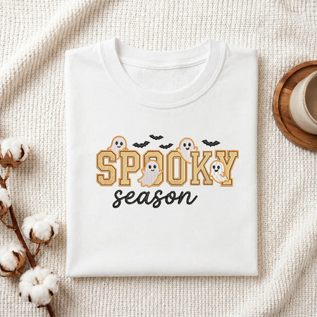 A folded white t-shirt with a 'Spooky Season' graphic print, styled in a flat-lay on a textured cream fabric with cotton stems and a coffee cup.
