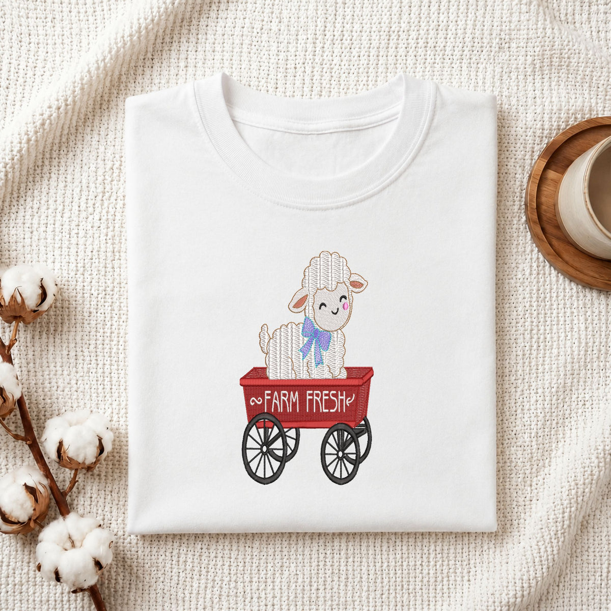 Folded white cotton t-shirt mockup featuring a cute "Farm Fresh" sheep machine embroidery design, styled with cotton stems and a wooden cup.   