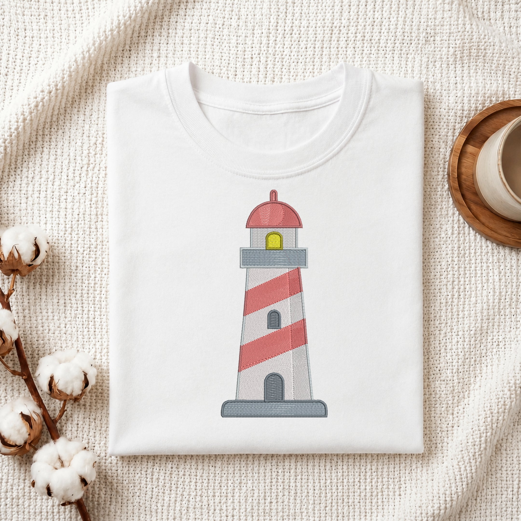 A folded white T-shirt with a lighthouse graphic printed on the front, styled on a textured cream fabric with cotton stems and a wooden cup in the background.