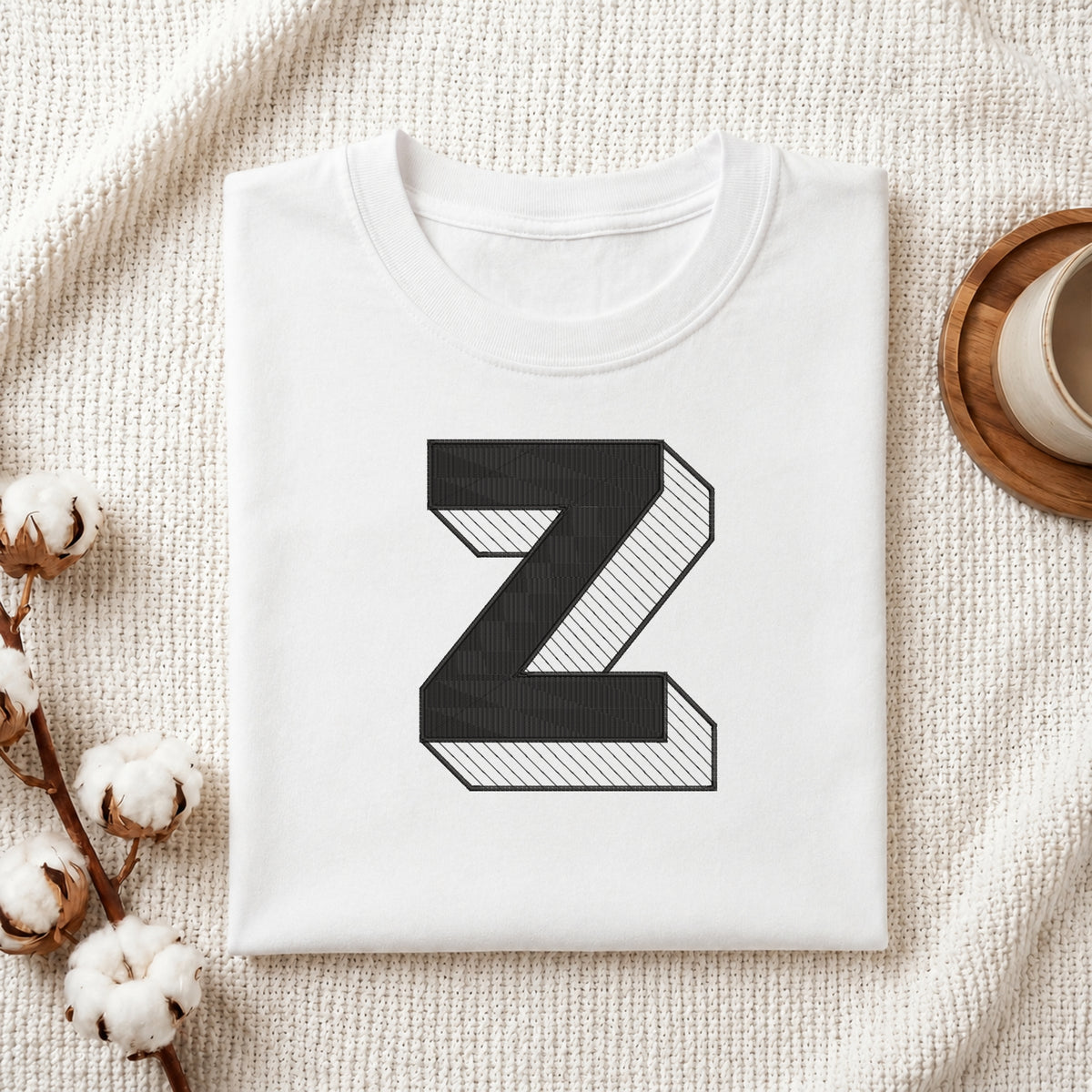 A folded white t-shirt with a large black 3D-style letter 'Z' printed on the front, styled on a textured cream fabric background with cotton bolls and a cup of coffee.