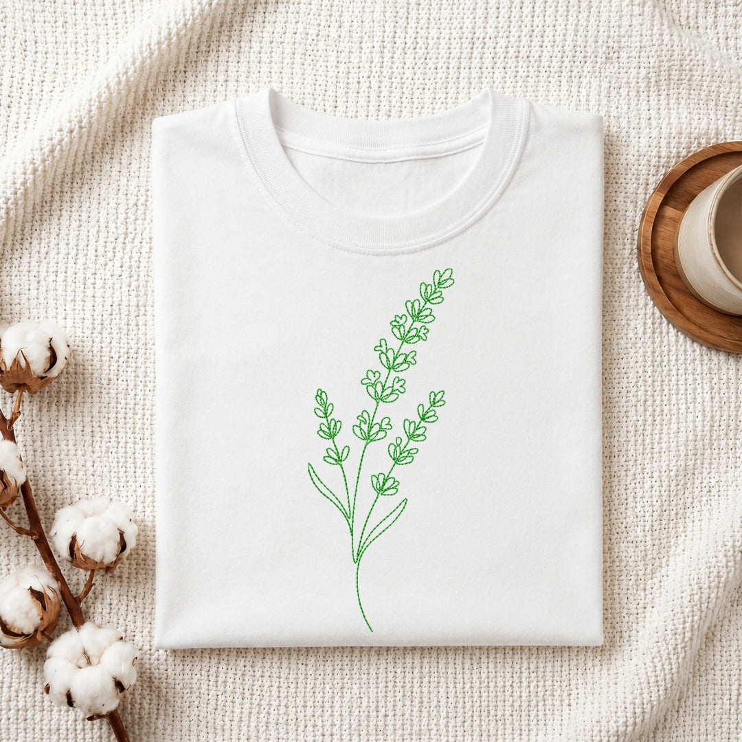 A white folded t-shirt featuring a green, line-art embroidered lavender design, presented on a textured white fabric with cotton sprigs and a wooden coaster.