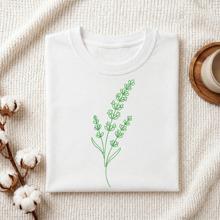 A white folded t-shirt featuring a green, line-art embroidered lavender design, presented on a textured white fabric with cotton sprigs and a wooden coaster.