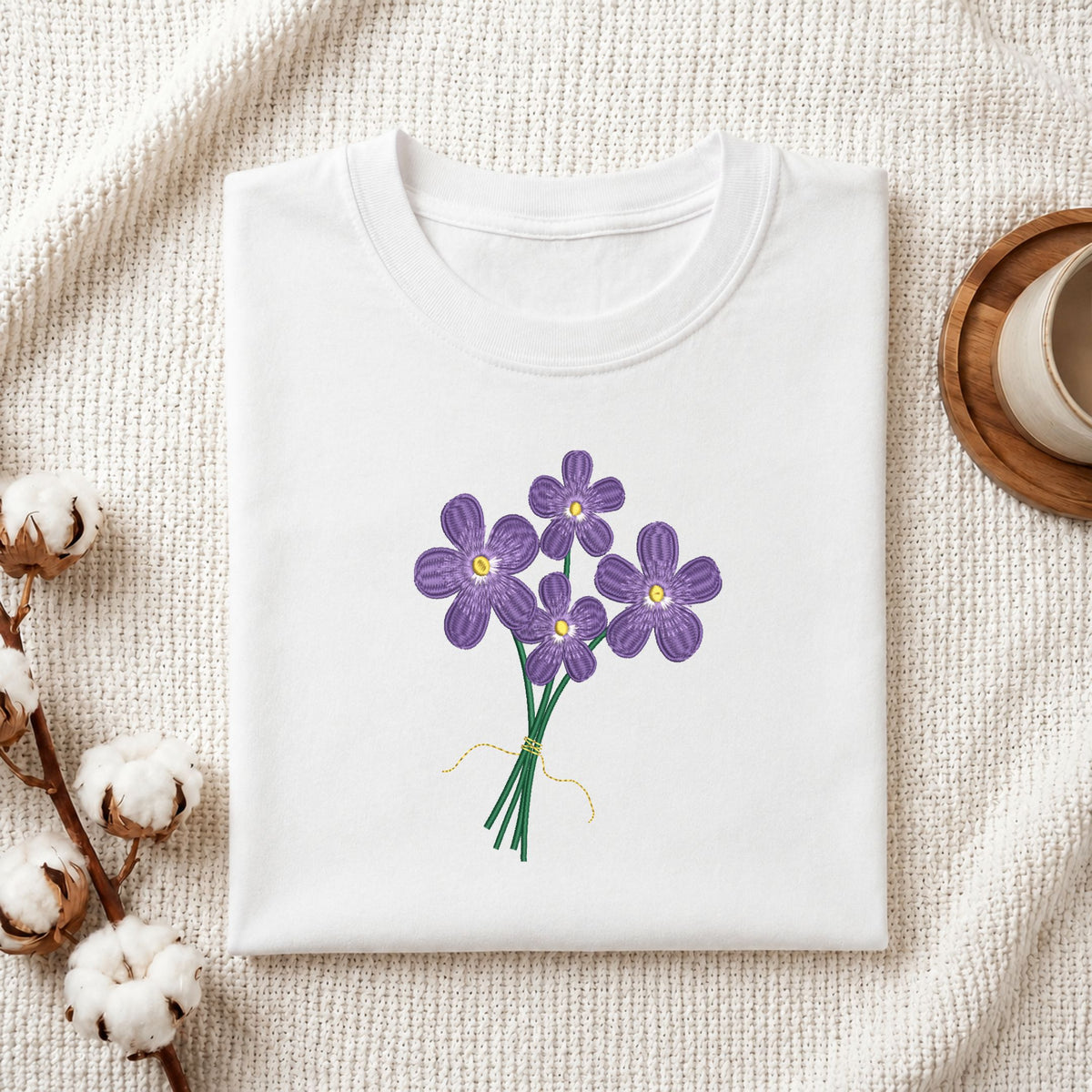 Folded white cotton t-shirt mockup featuring a purple flowers bouquet machine embroidery design, styled with cotton stems and a wooden cup