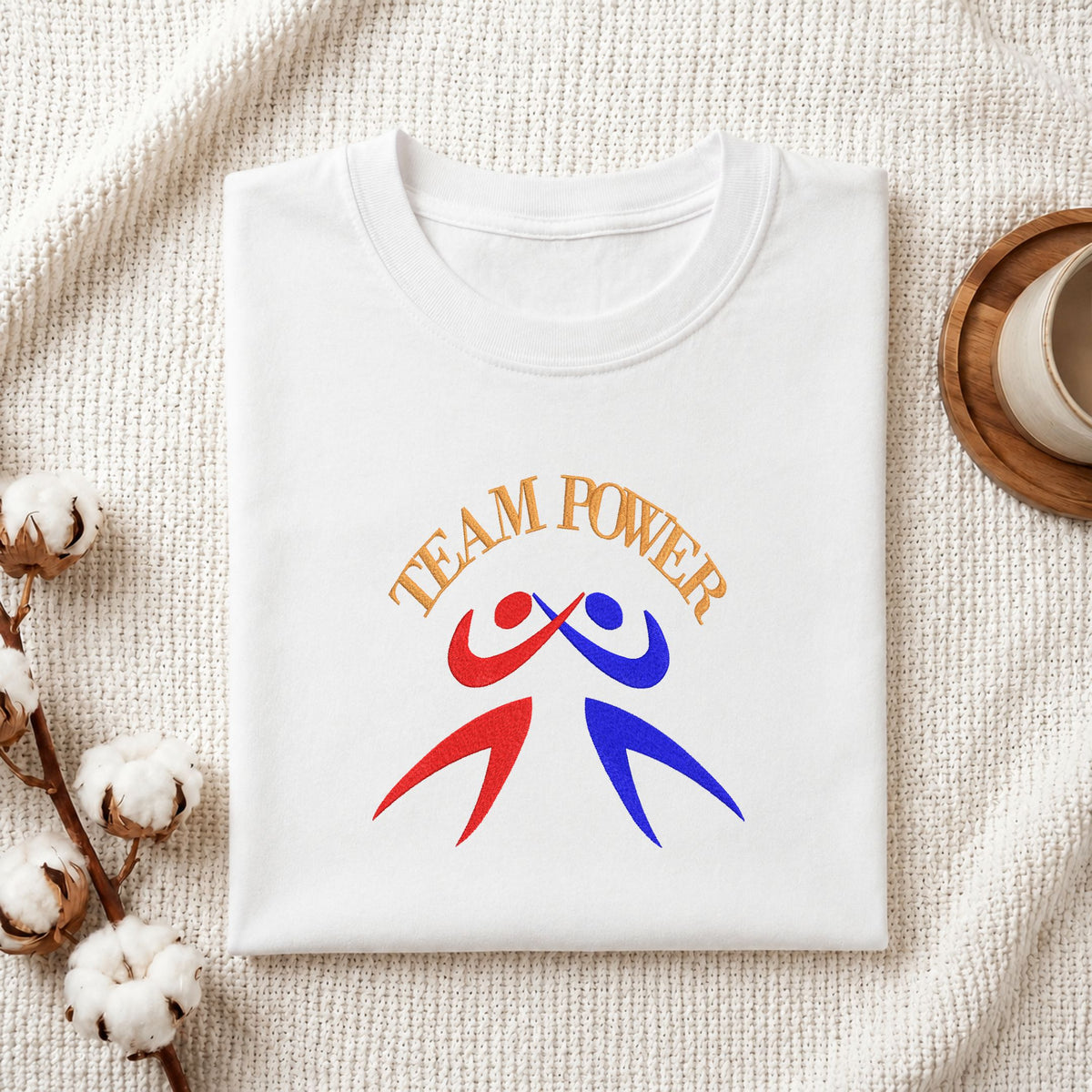 Folded white cotton t-shirt mockup featuring a "Team Power" machine embroidery design, styled with cotton stems and a wooden cup