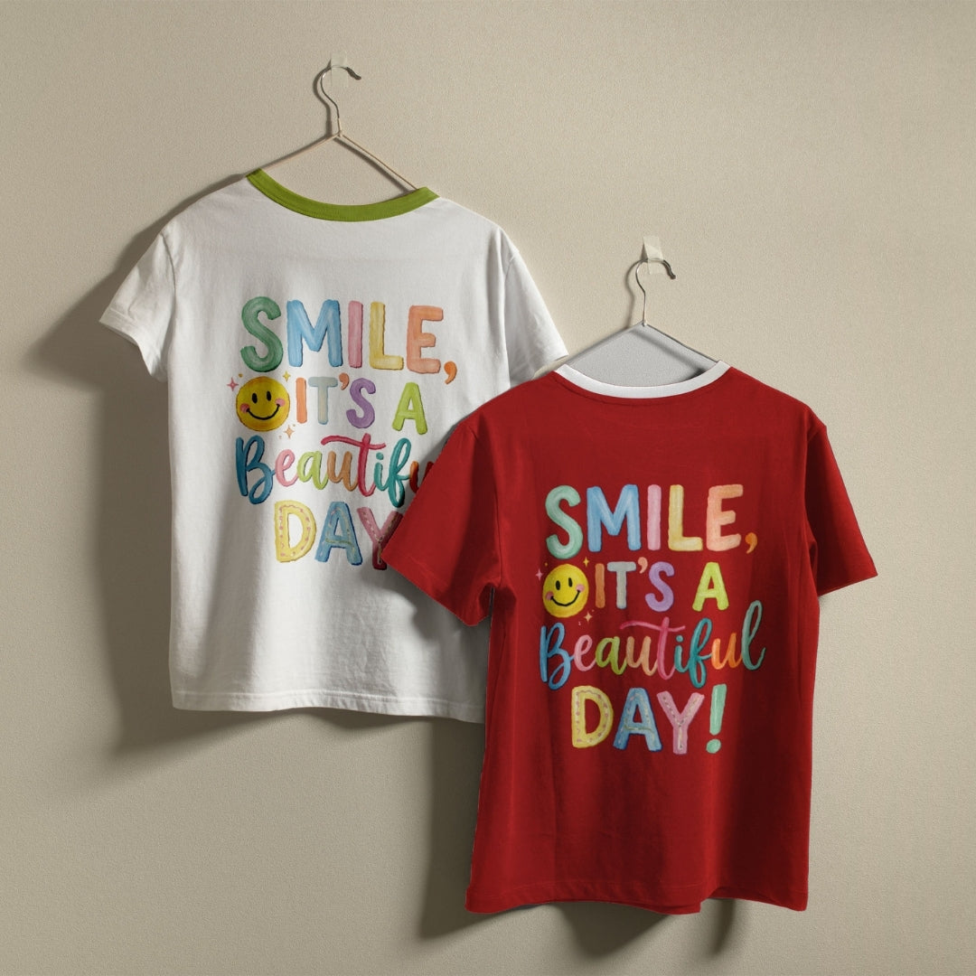 Two children's short-sleeve t-shirts hanging on hangers against a plain beige wall. The front shirt is bright red with a white collar and colorful text reading 'SMILE, IT'S A Beautiful DAY!' with a yellow smiley face.