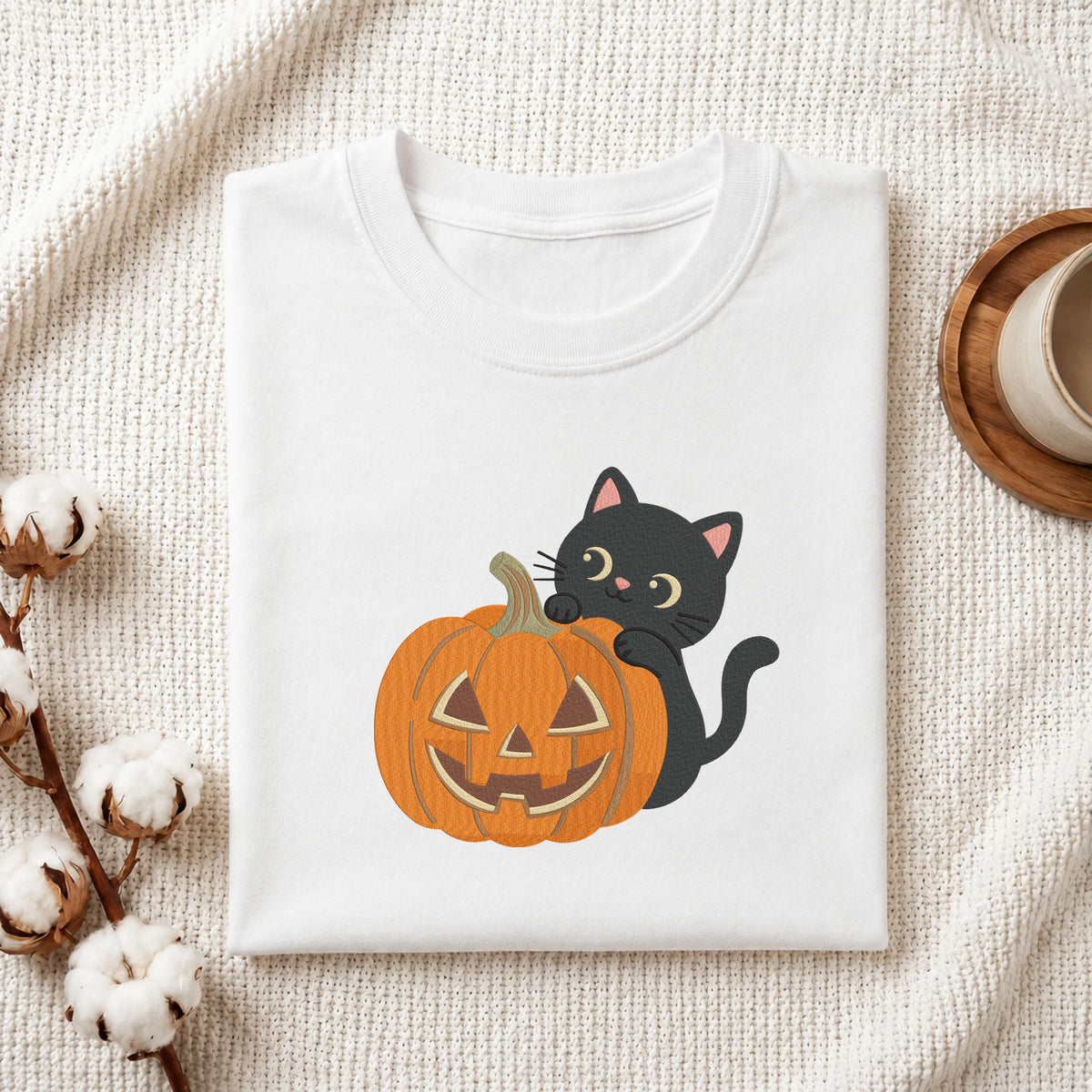 Folded white cotton t-shirt mockup featuring a cute black cat peeking behind a jack-o-lantern pumpkin machine embroidery design, styled with cotton stems and a wooden cup