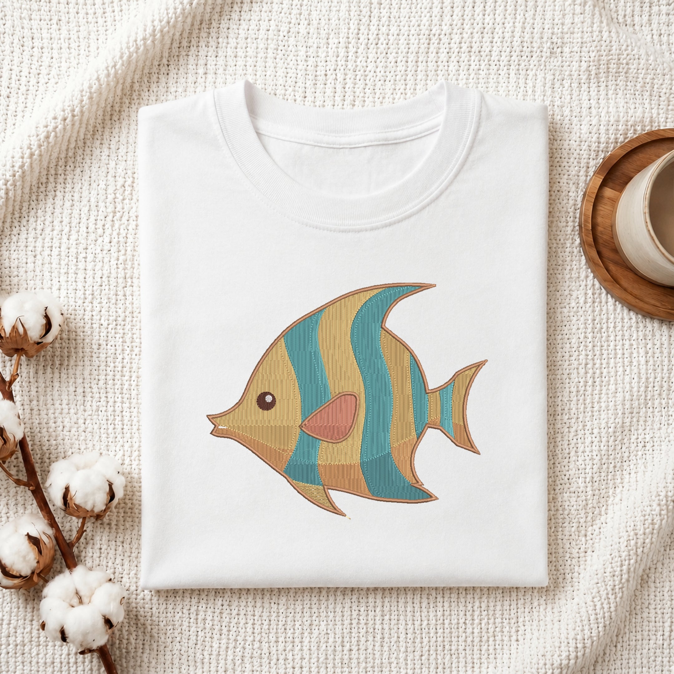 A folded white crewneck t-shirt with a colorful embroidered fish design, styled on a textured cream fabric with cotton bolls and a wooden cup.