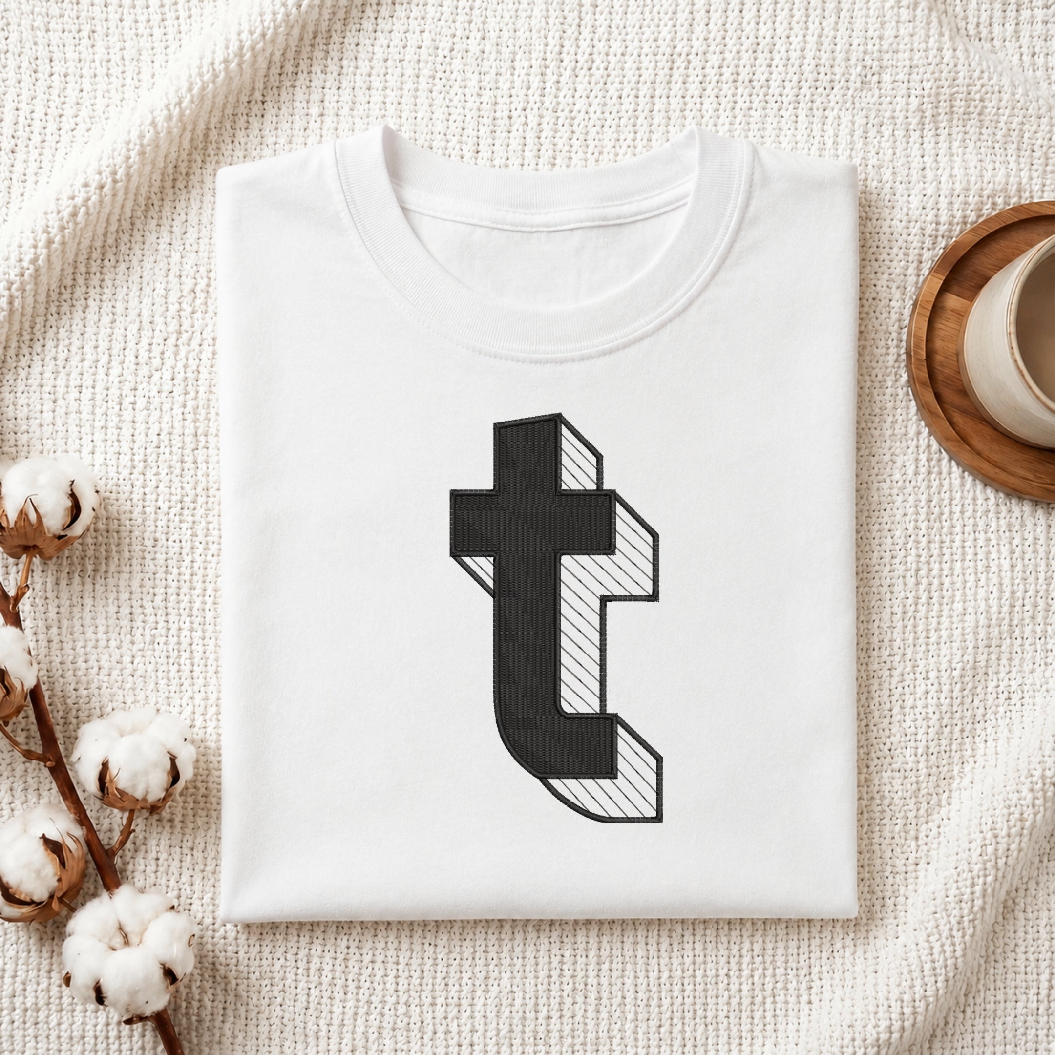 A folded white t-shirt with a black 3D-effect letter 't' graphic printed on the front, styled on a textured cream blanket with cotton bolls and a wooden cup in the background.