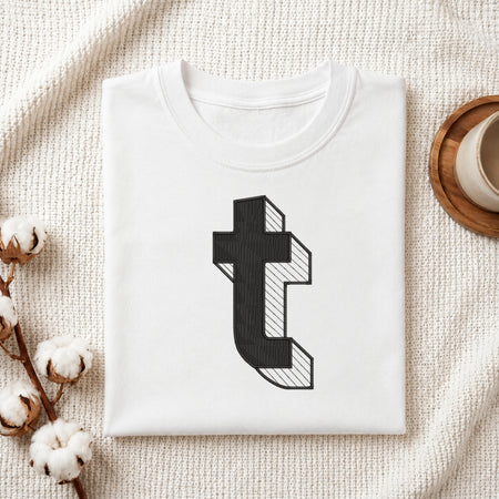 A folded white t-shirt with a black 3D-effect letter 't' graphic printed on the front, styled on a textured cream blanket with cotton bolls and a wooden cup in the background.