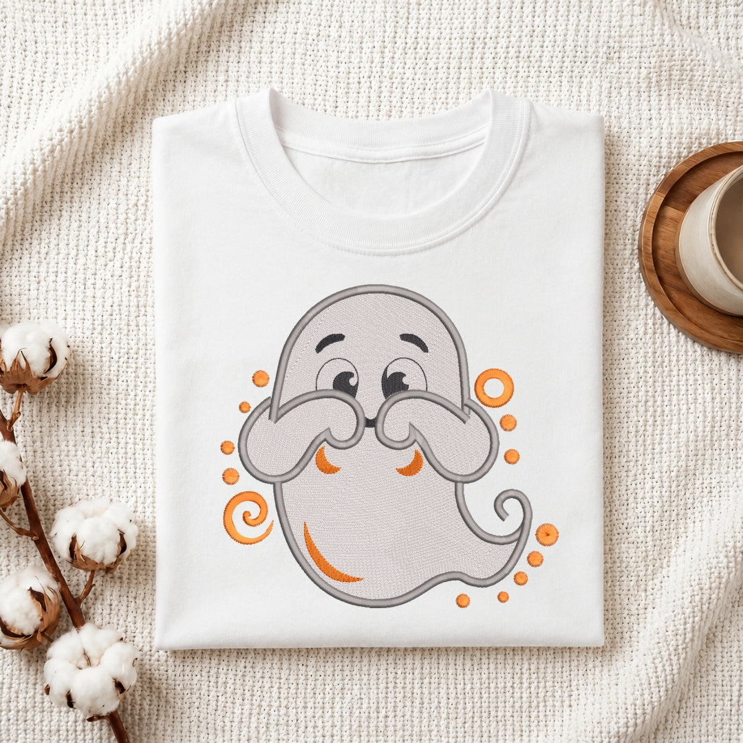 A folded white T-shirt with a cute cartoon ghost graphic in gray and orange, styled on a textured cream fabric with cotton stems and a small cup in the corner.