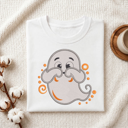 A folded white T-shirt with a cute cartoon ghost graphic in gray and orange, styled on a textured cream fabric with cotton stems and a small cup in the corner.