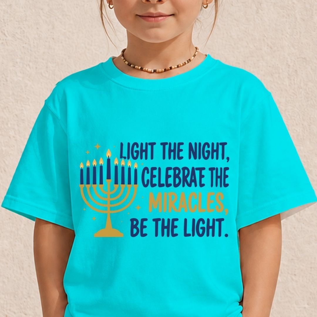 A child wearing a bright turquoise T-shirt with a printed graphic and text celebrating Hanukkah. The graphic features a yellow menorah and the text 'LIGHT THE NIGHT, CELEBRATE THE MIRACLES, BE THE LIGHT.