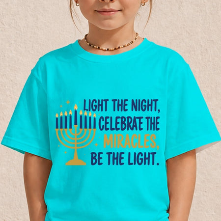 A child wearing a bright turquoise T-shirt with a printed graphic and text celebrating Hanukkah. The graphic features a yellow menorah and the text 'LIGHT THE NIGHT, CELEBRATE THE MIRACLES, BE THE LIGHT.