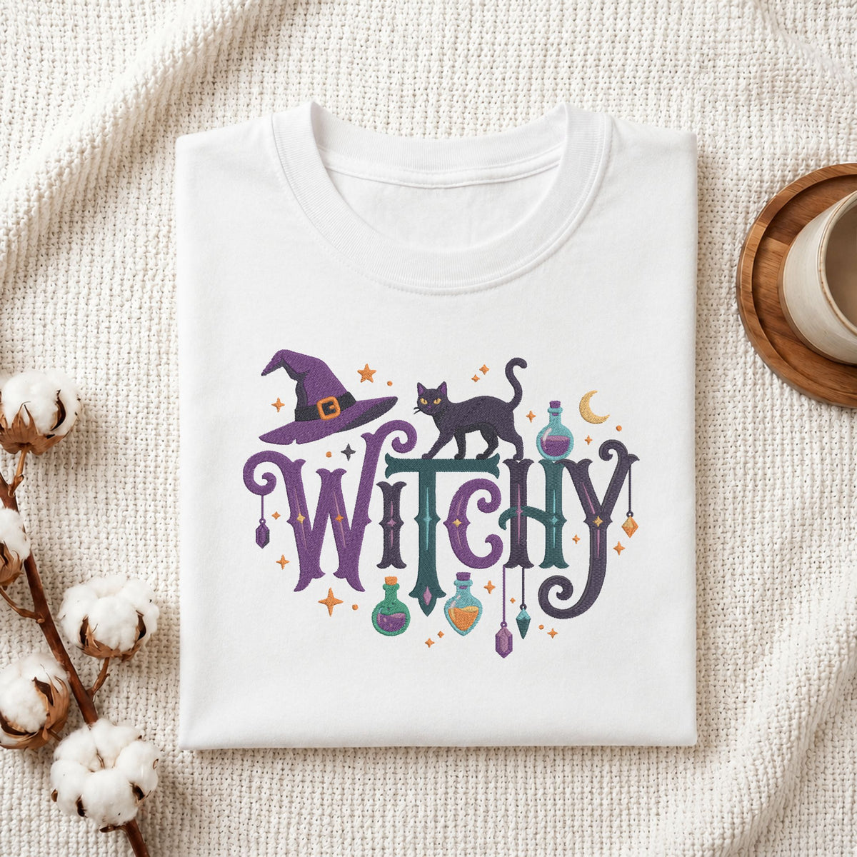 A folded white t-shirt with a colorful 'Witchy' graphic print featuring a witch hat, black cat, potion bottles, and stars, styled on a textured cream fabric with cotton bolls and a coffee cup.