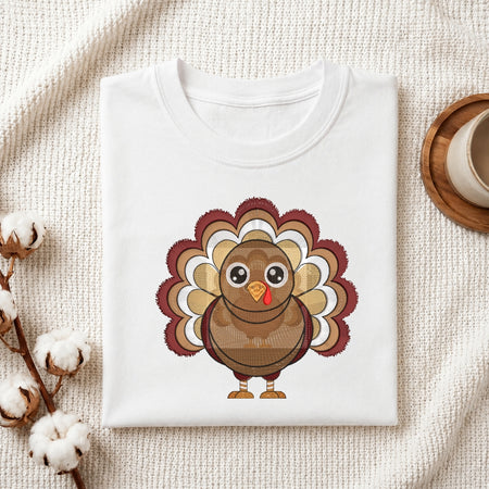 A folded white crewneck t-shirt featuring a cartoon turkey graphic, styled in a flat-lay on a textured cream fabric with cotton stems and a wooden cup.