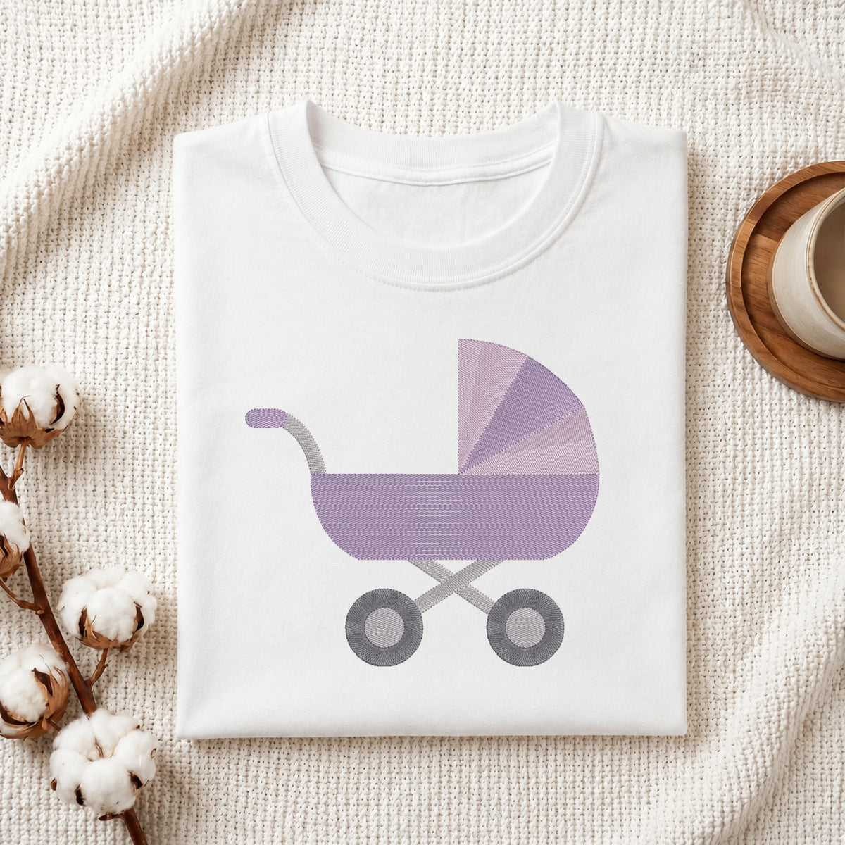 A folded white baby t-shirt with a purple and gray baby stroller graphic, styled on a textured cream blanket with cotton bolls and a wooden cup.