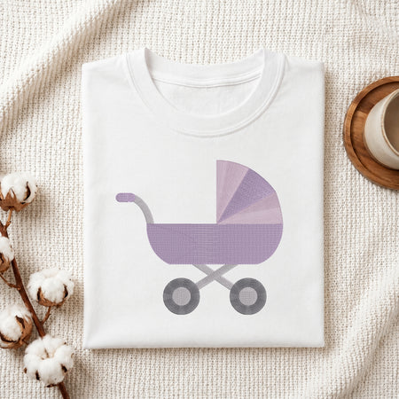 A folded white baby t-shirt with a purple and gray baby stroller graphic, styled on a textured cream blanket with cotton bolls and a wooden cup.