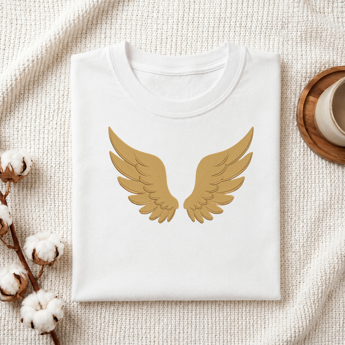 A folded white crewneck t-shirt with a gold angel wings graphic on the front, styled on a textured cream fabric with cotton bolls and a wooden cup in the background.