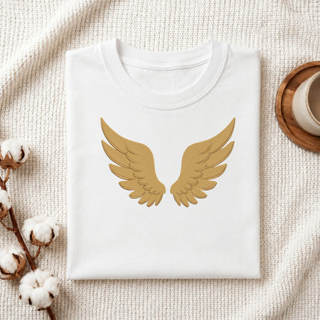 A folded white crewneck t-shirt with a gold angel wings graphic on the front, styled on a textured cream fabric with cotton bolls and a wooden cup in the background.