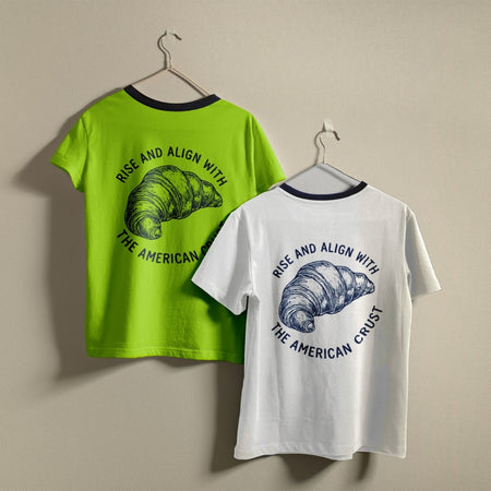 Two t-shirts hanging on a wall, one bright lime green and one white, both featuring the circular retro "Rise and Align with the American Crust" croissant design.
