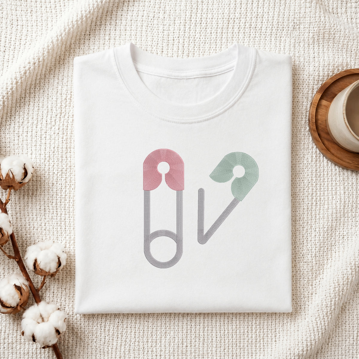 A folded white crewneck t-shirt with a graphic of two safety pins in pink, green, and gray, styled on a textured cream knit blanket with cotton bolls and a wooden cup in the background.