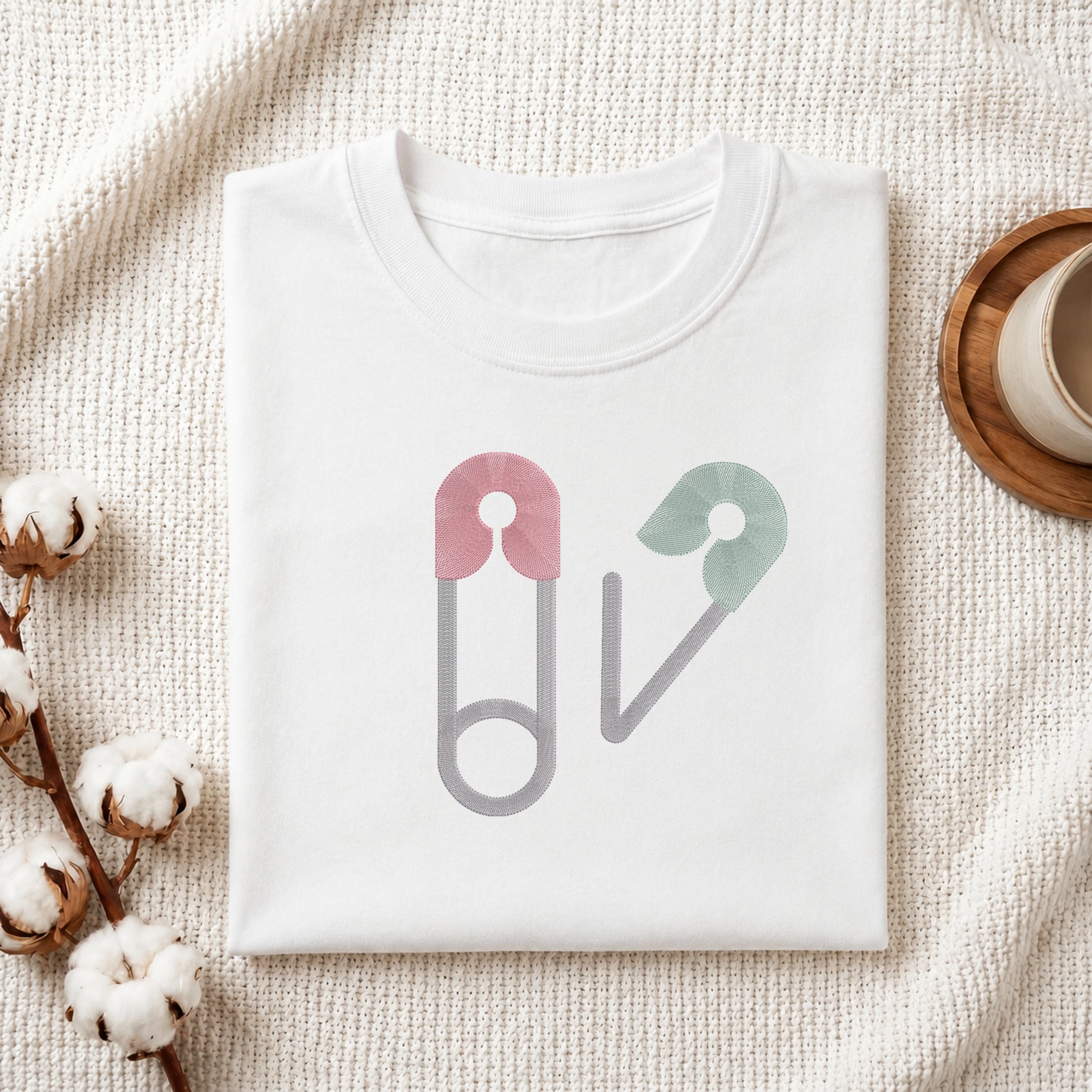 A folded white crewneck t-shirt with a graphic of two safety pins in pink, green, and gray, styled on a textured cream knit blanket with cotton bolls and a wooden cup in the background.