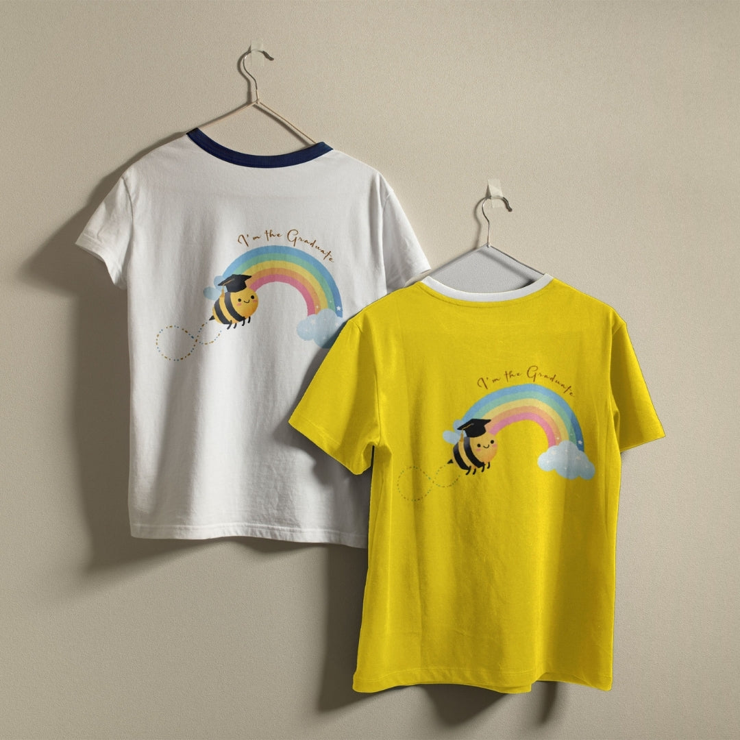 Two children's ringer t-shirts hanging on hangers against a plain beige wall. One shirt is white with a navy collar and features a bee wearing a graduation cap under a rainbow graphic with the text 'No more Graduals!'.