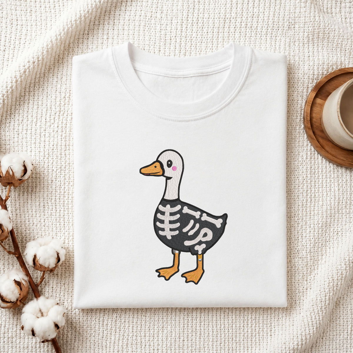 Folded white cotton t-shirt mockup featuring a cute skeleton duck Halloween machine embroidery design, styled with cotton stems and a wooden cup   