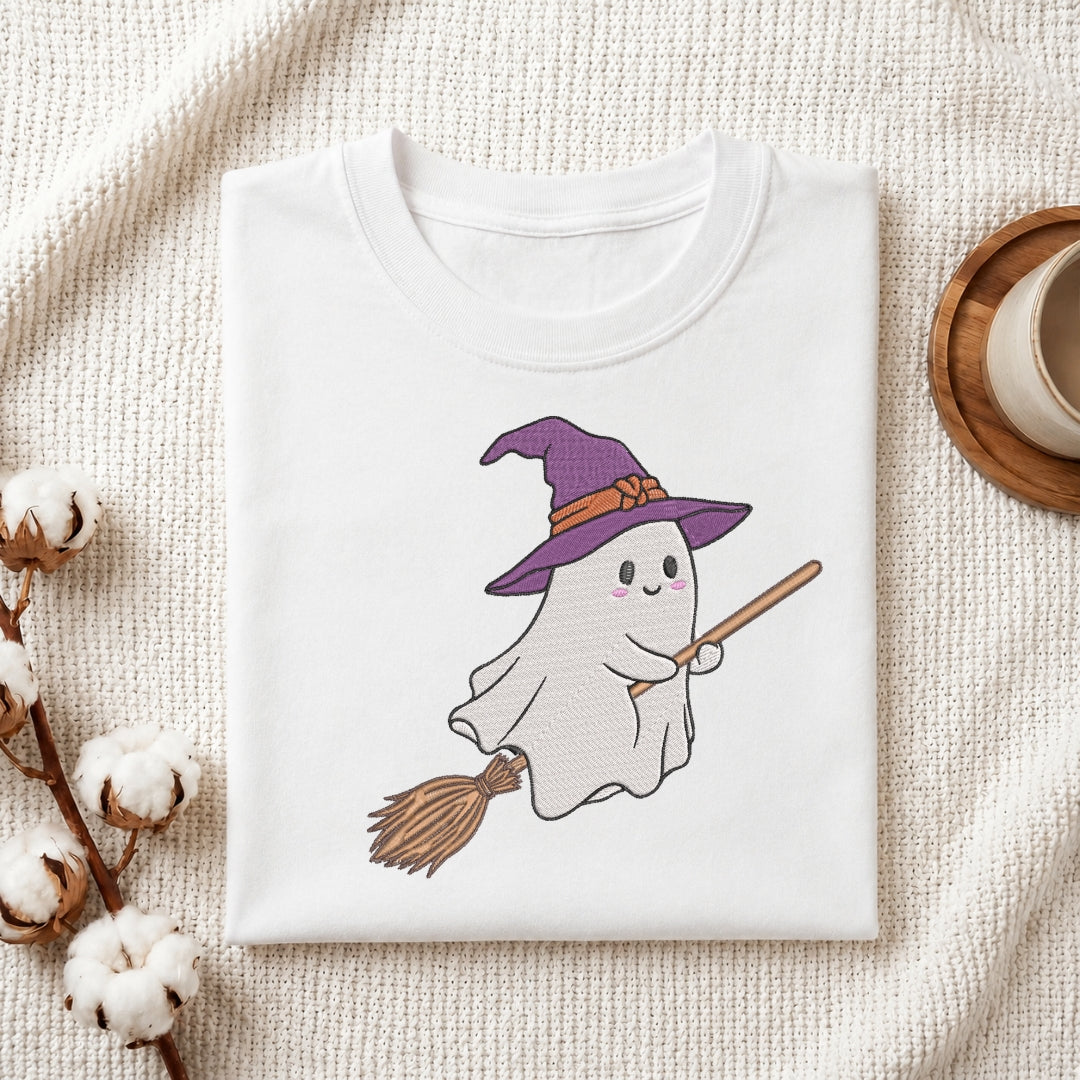 A folded white T-shirt with a cute embroidered-style graphic of a smiling ghost wearing a purple witch hat and holding a broomstick, displayed on a textured cream fabric with cotton stems and a small cup in the background.